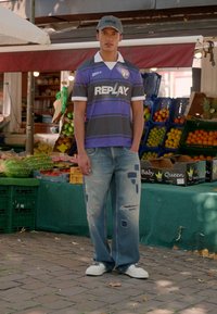Young man wearing a baseball cap, soccer jersey, patched jeans, and sneakers stands in front of a fruit market stall with assorted fruits.