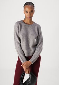 Suéter gris de punto con cuello redondo, puños y cinturilla acanalados, combinado con pantalones de pana burdeos y un bolso negro.