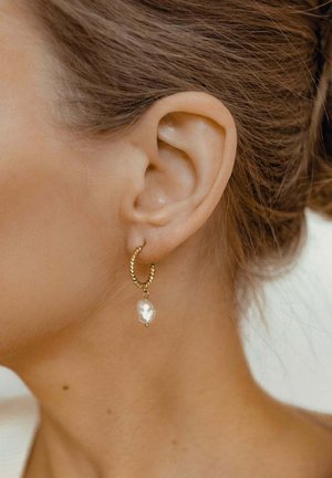 Boucle d'oreille en or avec un design torsadé, ornée d'une perle blanche irrégulière suspendue. Vue en gros plan mettant en valeur le lobe de l'oreille.