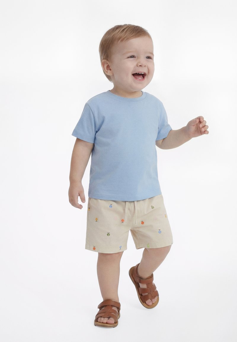Νήπιο που φοράει ανοιχτό μπλε μπλουζάκι, μπεζ σορτς με μικρά πολύχρωμα σχέδια φρούτων και καφέ σανδάλια, περπατάει και χαμογελά σε λευκό φόντο.
