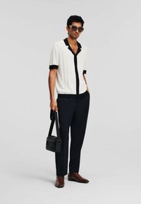 Witte shirt met korte mouwen en zwarte accenten, gecombineerd met donkere op maat gemaakte broeken en bruine schoenen; ook met een zonnebril en een kleine zwarte tas.