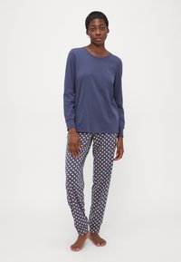 Kvinna som står barfota iförd en enkel långärmad blå topp och mönstrade blå och vita pyjamasbyxor mot en vit bakgrund.
