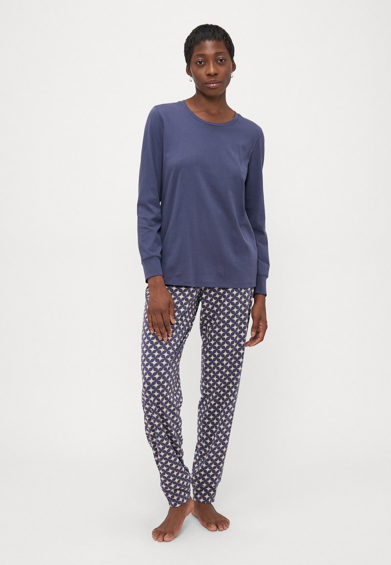 Vrouw staat blootsvoets in een eenvoudige blauwe longsleeve top en een patroon van blauw en witte pyjamabroek tegen een witte achtergrond.