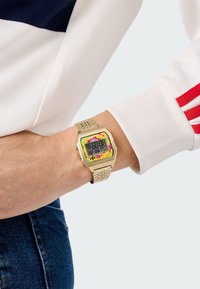 Digital klocka med ett guldmetallarmband, som har en rektangulär urtavla, klara färgaccenter runt urtavlan och digital visning.