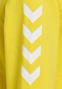 Gul tröja med texturerad yta, prydd med ett vitt chevronmönster på ärmen. Materialet är en bomullsblandning för komfort.