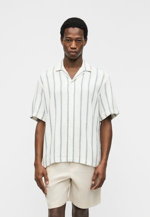 Άνδρας που φοράει λευκό μπλουζάκι με κοντά μανίκια και κάθετες ρίγες, καθώς και ανοιχτό μπεζ σορτς, στέκεται μπροστά σε απλό φόντο.