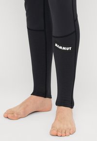 Juoda kojinė, pagaminta iš tekstūruoto audinio, su prigludusia forma, turinti matomą baltą "MAMMUT" logotipą šone. Atvira ties kulkšna.