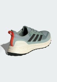 Behárová obuv s mätovo zeleným zvrškom, čiernymi pruhmi, oranžovou pútkom na päte a škvrnitou krémovou podrážkou. Textúrovaná sieťovina a podporný dizajn.