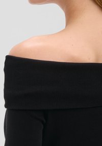 Primo piano di una spalla e collo che indossano una maglia nera a spalle scoperte in tessuto a coste su uno sfondo grigio chiaro uniforme.