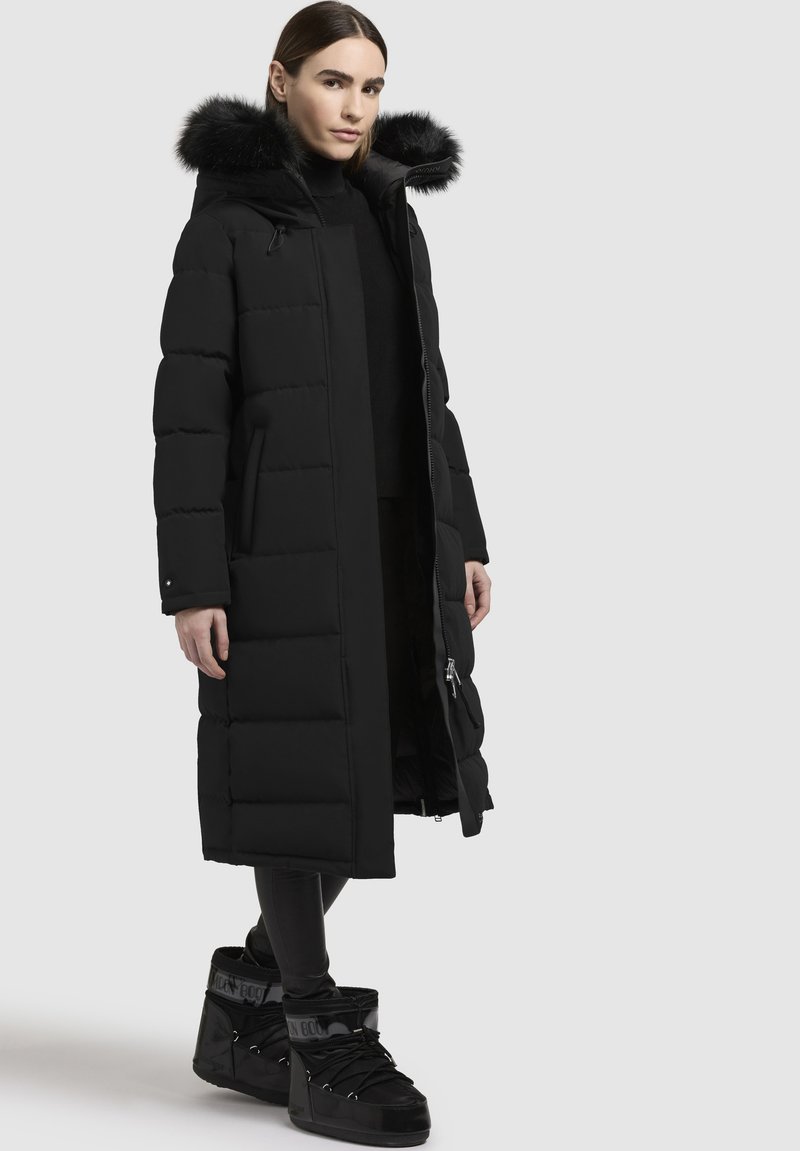 Dlouhý černý péřový kabát s kožešinovou kapucí, se zapínáním na zip, prošívaným designem a bočními kapsami; nošený s černými robustními kotníkovými botami.