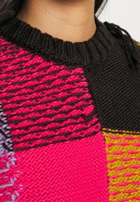 Multikleurige gebreide trui met opvallende patchwork in roze, zwart en geel, voorzien van textuurpatronen. Ribbelige zwarte halslijn.