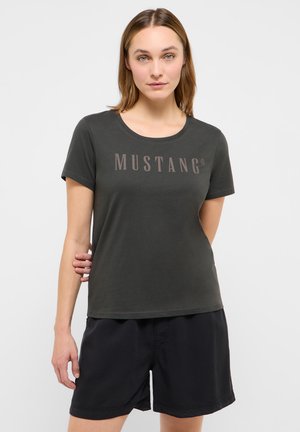 Frau steht mit den Händen im Rücken, trägt ein dunkelgraues T-Shirt mit Mustang-Logo und schwarze Shorts vor einem schlichten hellen Hintergrund.