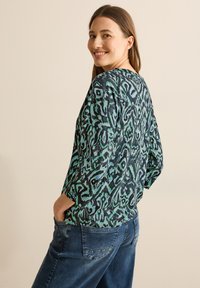 Haut à manches longues coloré avec un motif vert et marine, présentant une coupe décontractée et un col rond. Porté avec un jean en denim ample.