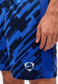 Близък план на ръка, почиваща върху спортни къси панталони Nike с синьо и черно шарка, с бяло лого на Nike близо до подгъва.