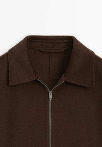 Brun zip-up jakke med spids krave, blød tekstur og metal lynlås. Enkel design uden synlige mønstre eller dekorationer.
