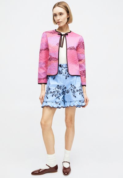 Γυναίκα που φοράει ροζ υφασμάτινο σακάκι με μαύρο περιθώριο, ανοιχτό μπλε κεντημένες σορτς με βολάν, λευκές κάλτσες και καφέ παπούτσια Mary Jane, όρθια.