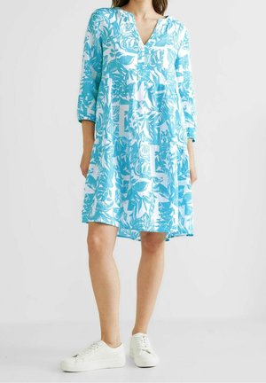 Femme portant une robe blanche ample longueur genou avec des imprimés botaniques bleus et des manches trois-quarts, associée à des baskets blanches.