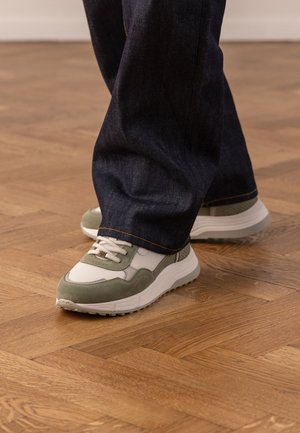 Persoon die witte en groene sneakers draagt met beige veters en donkerblauwe spijkerbroek, staand op een houten parketvloer.