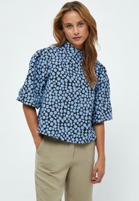 Blusa con stampa floreale e maniche corte a palloncino, caratterizzata da un colletto alto e uno sfondo nero con piccoli fiori blu. Tessuto morbido.