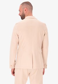 Blazer beige su misura con rever a scollo a V, apertura posteriore singola e polsini abbottonati, realizzato in tessuto liscio, che mostra una vista laterale.