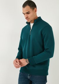 Pullover verde in pile con zip a metà, colletto alto, maniche lunghe e polsini a costine. Design casual con una texture liscia.