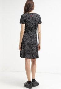 Schwarzes Kleid mit floralem Muster in Grautönen. Kurze Ärmel, taillierter Oberteil, ausgestellter Rock. Getragen mit schwarzen Plateauschuhen. Schlichter Hintergrund.