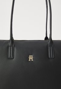 Sac à main en cuir noir avec deux poignées plates, arborant un emblème doré à l'avant. Texture lisse et fermeture éclair sur le dessus.