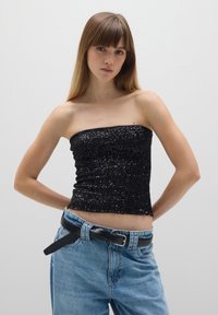 Top sin mangas de lentejuelas negras con una textura fruncida. Combinado con jeans de denim de cintura alta en un tono azul claro y un cinturón negro.
