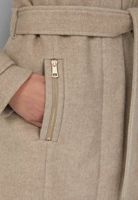 Manteau en mélange de laine beige avec une finition texturée, une ceinture à la taille et une poche zippée sur le côté avec des accessoires dorés.