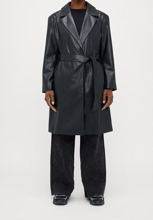 Personne portant un trench-coat en cuir synthétique noir ceinturé, un pantalon noir à jambes larges avec des paillettes subtiles, et des baskets noires sur un fond blanc.