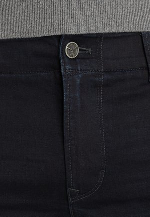 Temno modri jeans z gladko teksturo, ki imajo kovinsko zapiranje s gumbom, kontrastno šivanje in ozko prileganje okoli pasu.