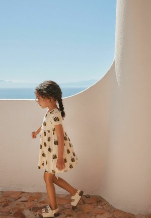 Young girl with black braided hair walks on stone floor wearing cream dress with bee prints and sandals by white curved wall and ocean view.