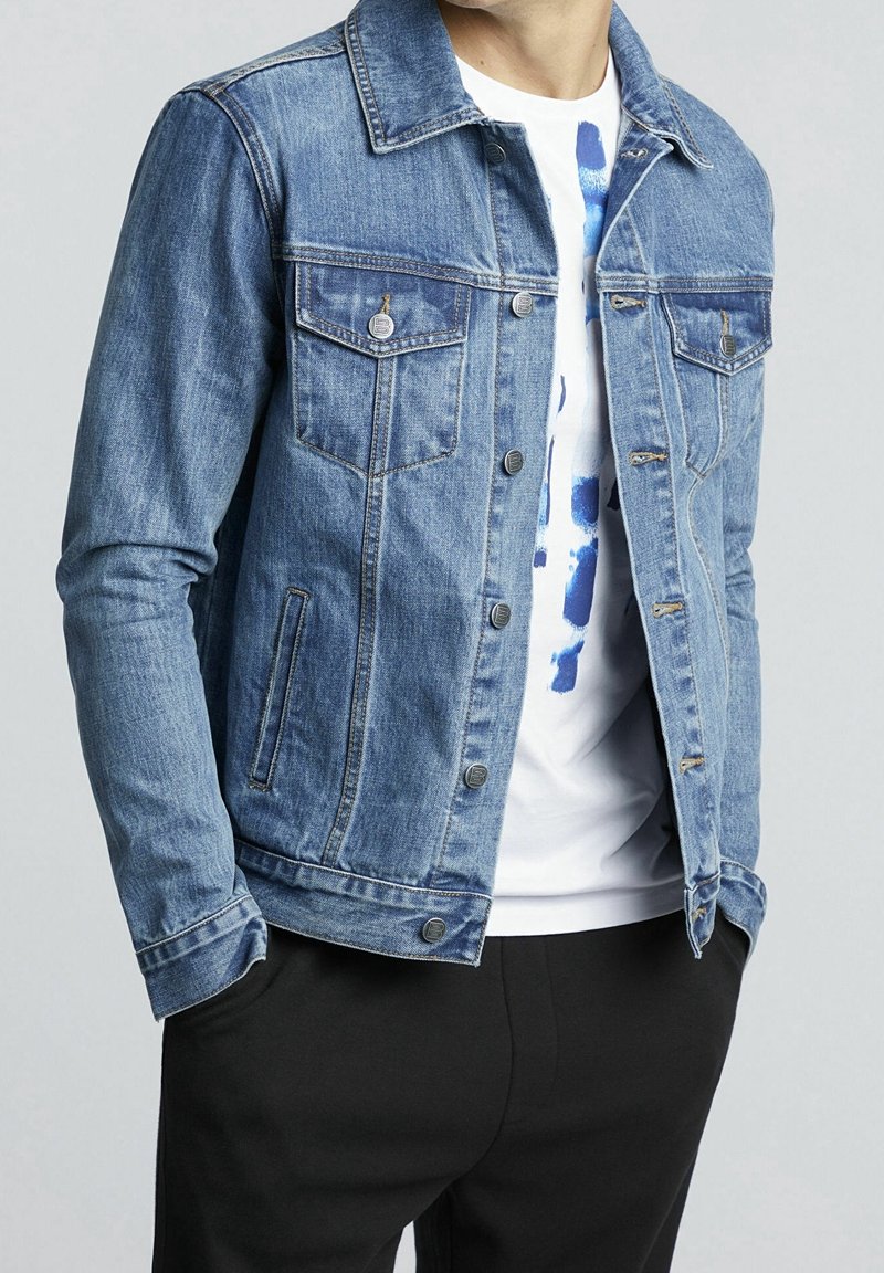 Muž v modré džínové bundě přes bílém tričku s modrým designem a černých kalhotách, s rukama v kapsách, na jednobarevném světlem pozadí.