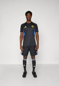 Μαύρη φανέλα ποδοσφαίρου με μπλε λεπτομέρειες και κίτρινο λογότυπο, συνοδευόμενη από μαύρο σορτς, μαύρες κάλτσες με λευκές ρίγες και μαύρα ποδοσφαιρικά παπούτσια με τάπες.