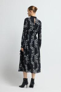 Schwarzes Kleid mit weißem Paisley-Muster, langen Ärmeln, geraffter Taille und einem Schlüsselloch-Detail am Rücken. Kombiniert mit schwarzen Ankle Boots.