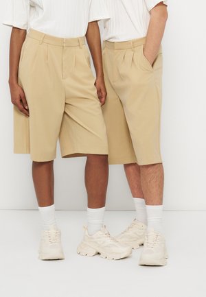 Dvaja ľudia oblečení v béžových kraťasoch dĺžky po kolená, bielych ponožkách, masívnych krémovo-bielych teniskách a bielych tričkách s krátkym rukávom, stoja vedľa seba.