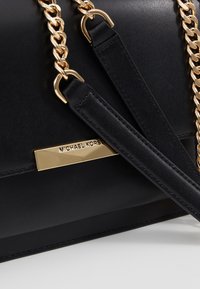 Černá kožená kabelka se zlatým řetízkovým popruhem, s kovovou destičkou s logem a hladkou texturou. Doplňky zahrnují nastavitelné kožené popruhy.