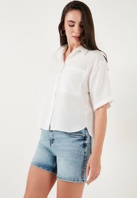 White linen short-sleeve button-up shirt with a chest pocket, paired with light blue denim shorts, featuring a fitted waist and rolled hems.