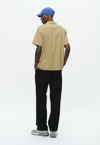 Chemise beige à manches courtes, pantalon noir et baskets grises. Une casquette bleue complète le look. Le tissu semble léger avec une texture lisse.