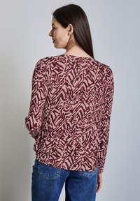 Langärmliges Oberteil in Kastanienbraun und Creme mit abstraktem Wirbelmuster, mit rundem Ausschnitt und lockerer Passform. Kombiniert mit blauen Jeans.