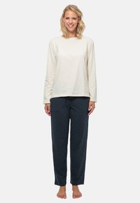 Ljus beige långärmad topp i kombination med mörkblå pyjamasbyxor med prickar. Tyget ser mjukt ut med en avslappnad passform. Barfota ställning.