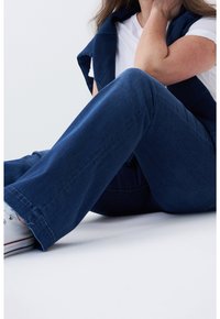 Personne assise par terre portant un jean bleu, des baskets blanches, un t-shirt blanc et un pull bleu marine posé sur les épaules.