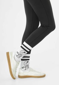 Weiße Crew-Socken mit schwarzen Streifen zeigen ein Snoopy-Graphic. Kombiniert mit schwarzen Leggings und weißen Sneakers mit einer Gummisohle.