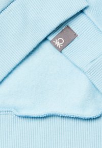 Světle modrý svetr s žebrovaným výstřihem a manžetami, vyrobený z měkké látky; má šedou visačku s bílým logem.