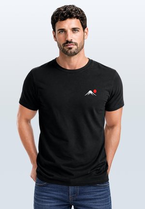 Man with short curly dark hair and beard wears black t-shirt with small white mountain and red sun design, standing against plain background.