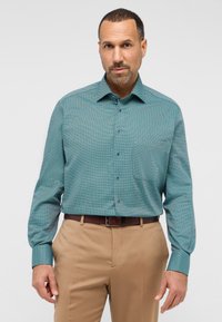 Tyrkis mønstret skjorte med lange ærmer, en brystlomme og blå knapper, parret med beige bukser. Glat stofstruktur.