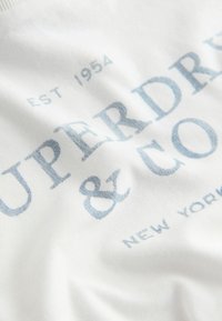 Balts kokvilnas T-krekls ar gaiši zilu drukātu tekstu. Teksts ietver "SUPERDRY & CO" ar "EST 1954" un "NEW YORK" detaļām. Gluda tekstūra.
