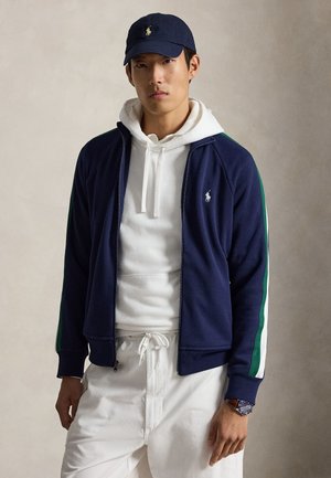 Giovane uomo che indossa un cappellino blu navy, una giacca con zip blu navy con strisce verdi e bianche, una felpa bianca con cappuccio e pantaloni bianchi con coulisse, in piedi davanti a uno sfondo semplice.
