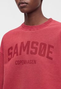 Gros plan d'une personne portant un sweat-shirt rouge avec "SAMSOE COPENHAGEN" imprimé sur le devant en lettres rouge foncé.