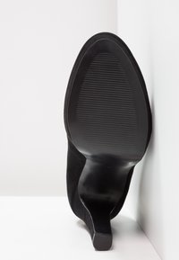 Černá semišová podešev vysokého podpatku, s hladkým povrchem, zakulacenou špičkou a texturovaným vzorem gripu, nakloněná proti bílému pozadí.